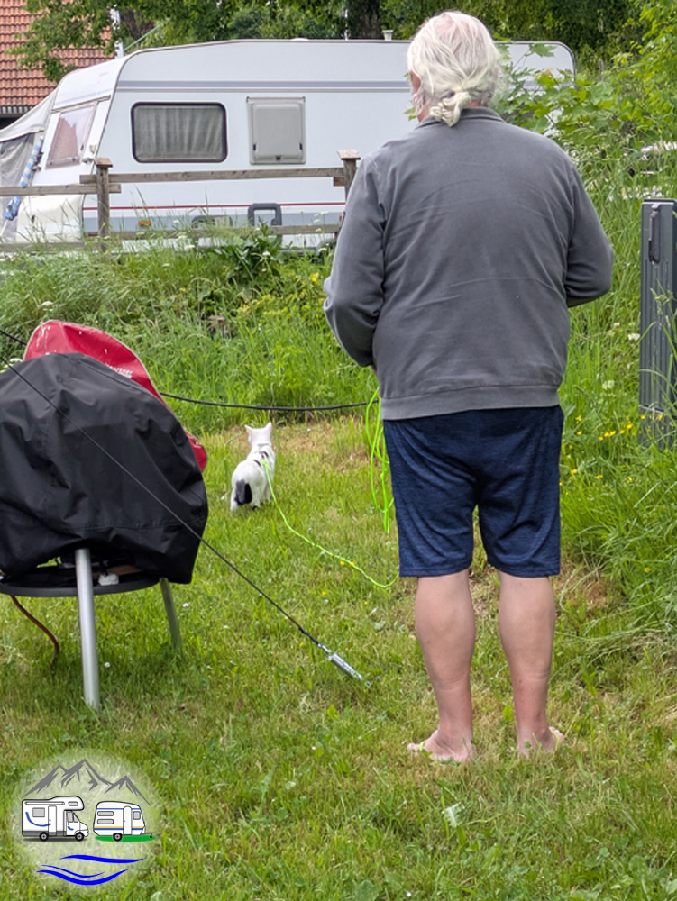 Eine Person steht mit dem Rücken zur Kamera und hält eine Katze an der Leine auf einer Wiese neben dem verhülltem Grill. Im Hintergrund ist ein Wohnwagen zu sehen.