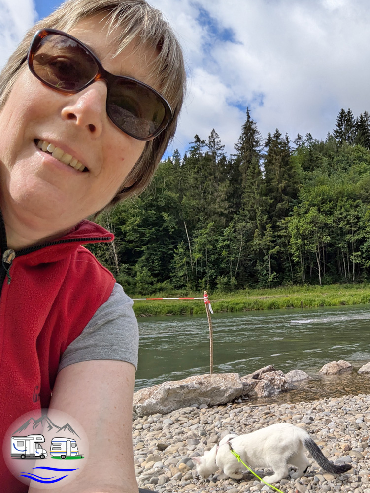 Eine Person mit Sonnenbrille macht ein Selfie am Flussufer, im Hintergrund schnüffelt eine weiße Katze an der Leine an den Ufersteinen.
