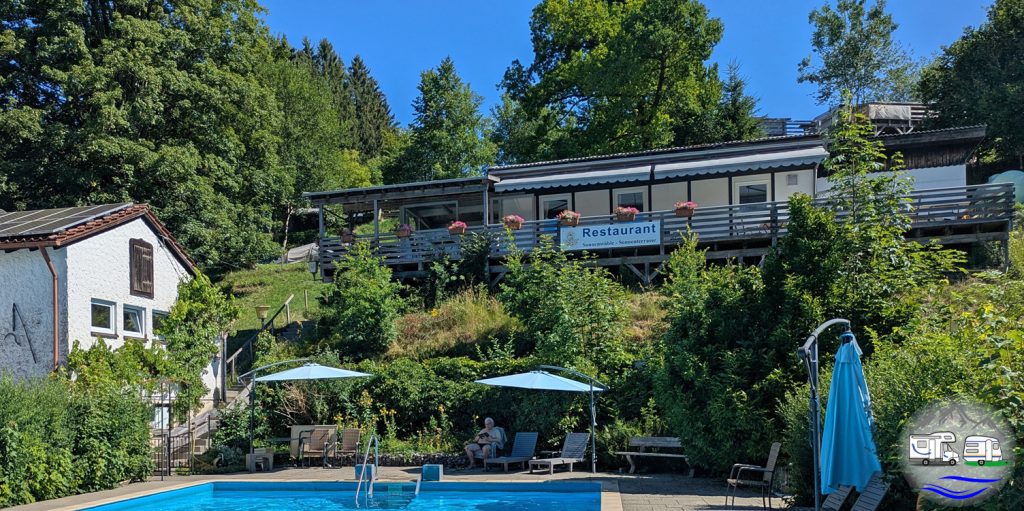 Restaurant mit Terrasse am Hang oberhalb eines Pools auf dem Campingplatz Sonnenbuckl, umgeben von Bäumen